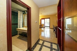 Un pasillo de hotel con suelo de baldosas de mármol, un marco de puerta de madera y vistas a un baño con pared de azulejos verdes y un lavabo blanco.