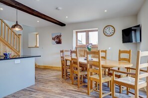 Un coin repas avec des chaises en bois et une table ronde, un escalier en bois, une horloge et un téléviseur fixé au mur.