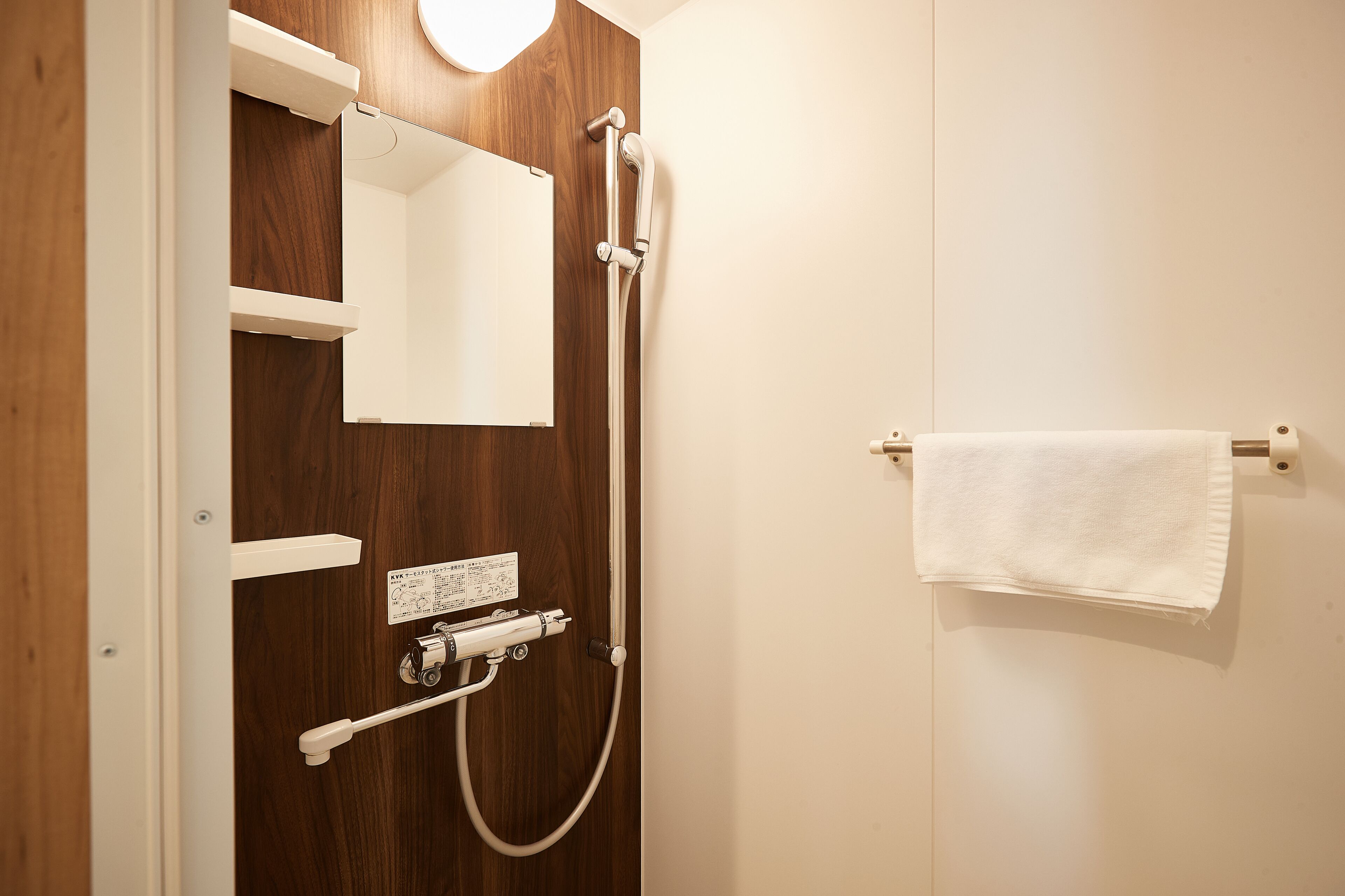A bathroom with a wooden door, a white towel hanging on a towel rack, a mirror, and a shower with a handheld showerhead.