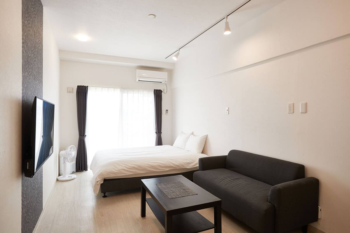 Quarto de hotel com cama, sofá escuro, mesa pequena, televisão e ar condicionado.