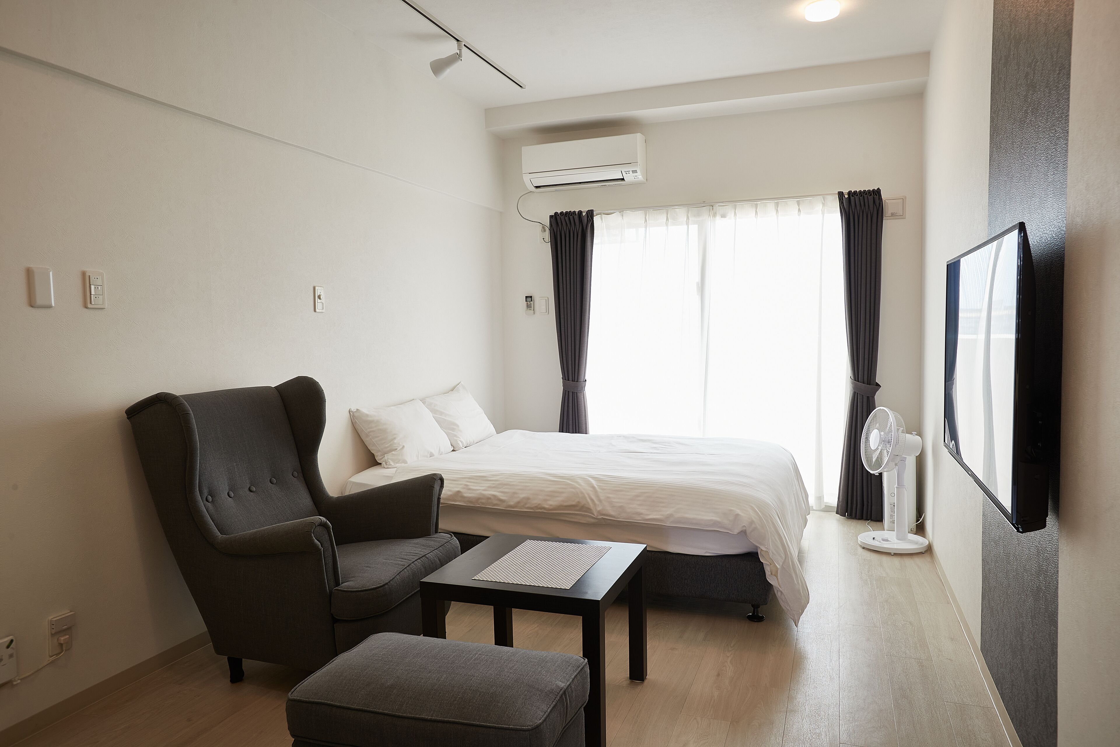 A hotel room with a bed, a chair, a table, an air conditioner, a fan, and a TV mounted on the wall.