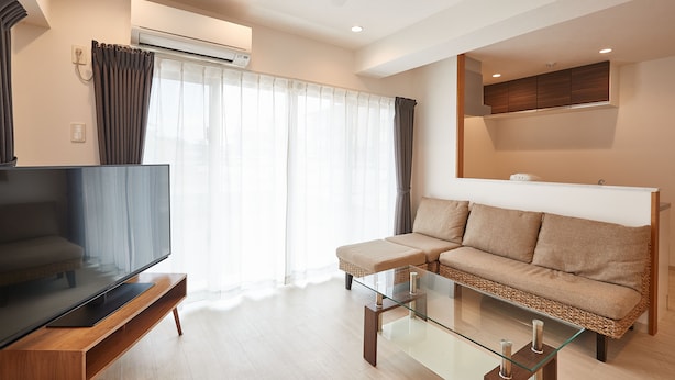 A modern living room with a flat-screen TV on a wooden stand, a glass coffee table, a beige sofa, and a ceiling fan. - New Normal Hotel in NAGO (Nago)