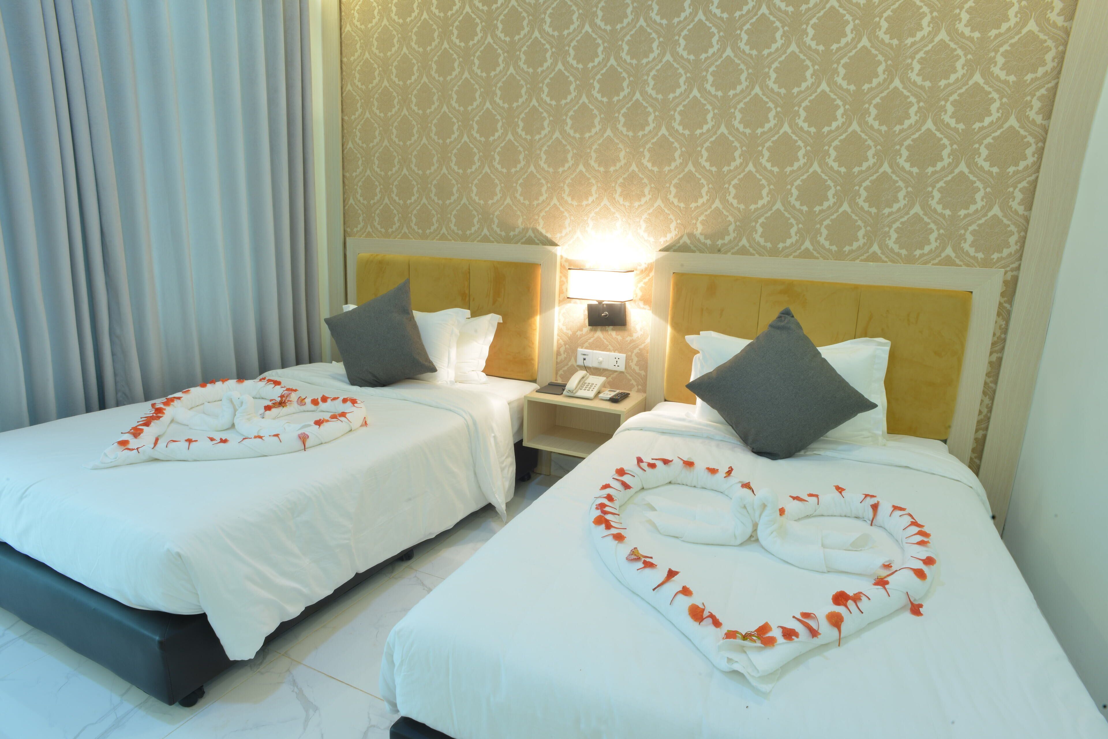 A hotel room with two beds, each with a heart-shaped towel arrangement and pillows. There is a nightstand with a lamp and a phone between the beds.