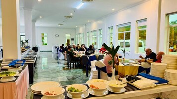 Una larga mesa de comedor con varias personas sentadas, rodeada de sillas blancas y cintas azules, en una habitación bien iluminada con ventanas grandes y piso de mármol.