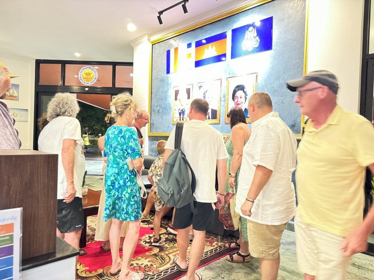 Een groep mensen staat in een hal met een receptiebalie, een kamerbreed tapijt en ingelijste foto's aan de muur.