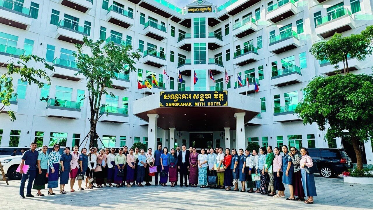 A group of people standing in front of the Sangkhak Mith Hotel, with multiple flags and a signboard displaying the hotel's name.