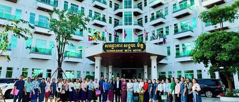 A group of people standing in front of the Sangkhak Mith Hotel, with multiple flags and a signboard displaying the hotel's name.
