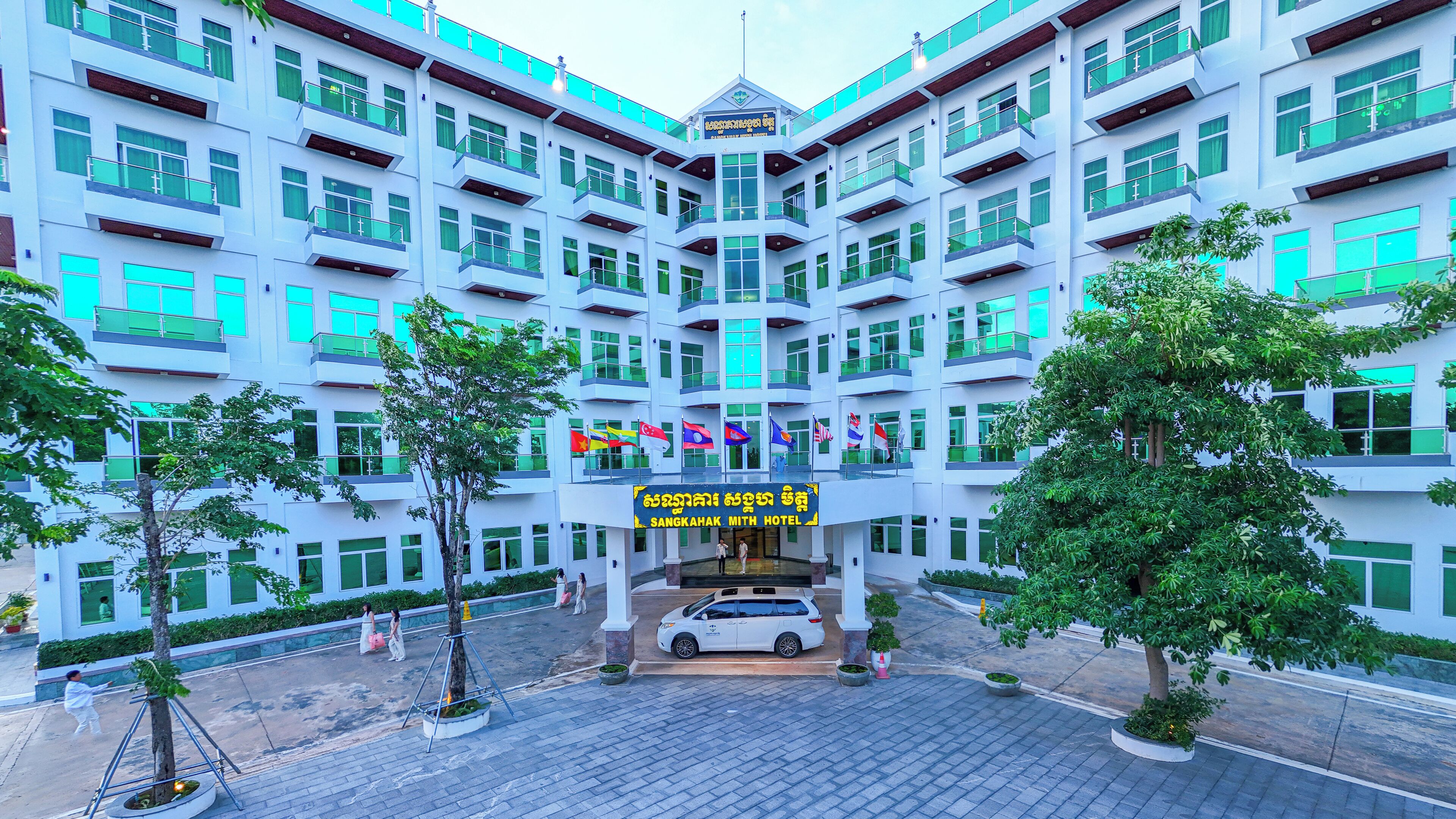 Ein mehrstöckiges Hotel mit zentralem Eingang, umgeben von Bäumen und Topfpflanzen.