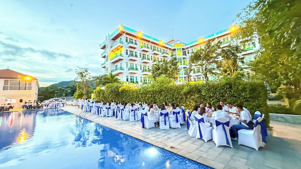 Zona junto a la piscina con sillas y mesas blancas, un edificio con balcones y un cielo despejado.