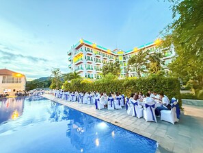 A poolside area with white chairs and tables, a building with balconies, and a clear sky.