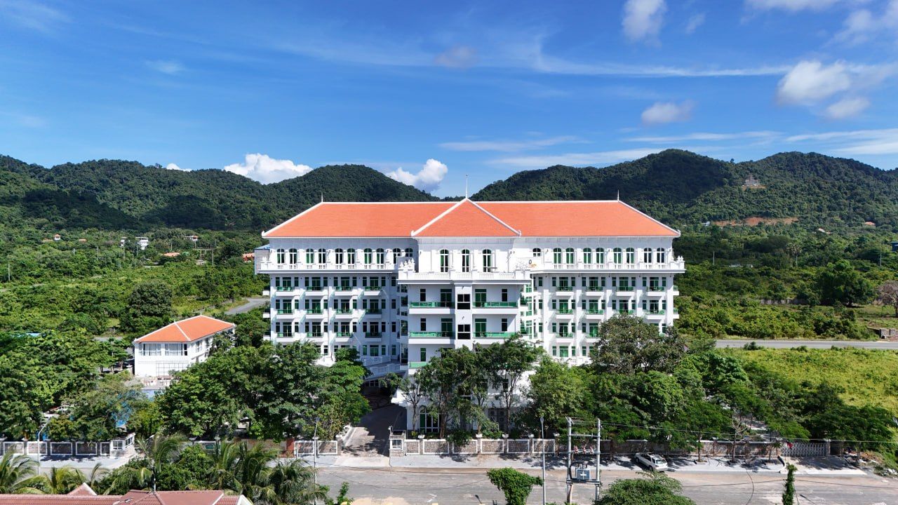 A large, multi-story building with a red roof, surrounded by lush greenery and hills.
