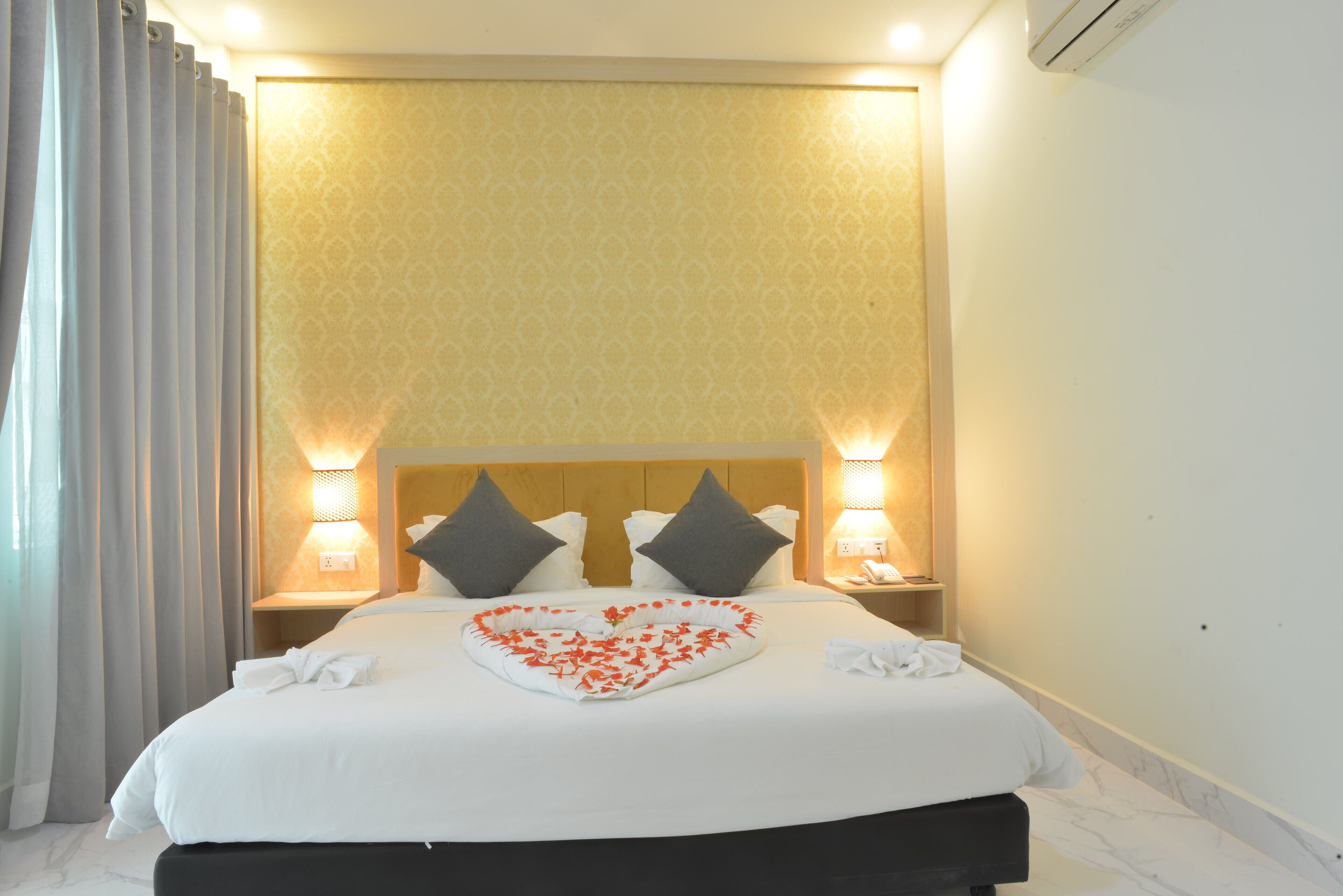 A hotel room with a neatly made bed, two bedside lamps, a headboard with two pillows, and a decorative heart-shaped arrangement on the bed.
