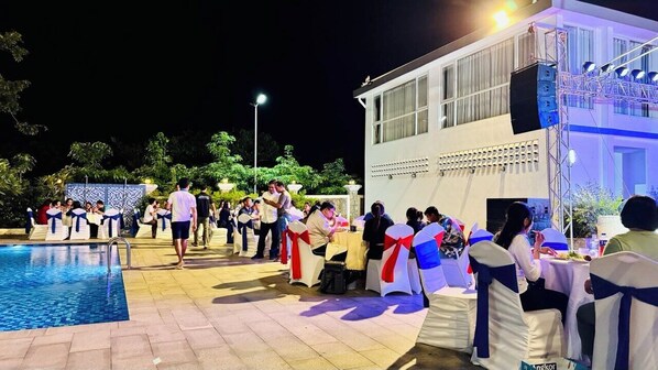 An outdoor event with people seated at tables, a pool, and a building with a balcony.