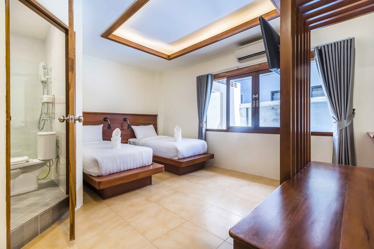 Quarto de hotel com duas camas, uma mesa de madeira, chuveiro e janela com cortinas.