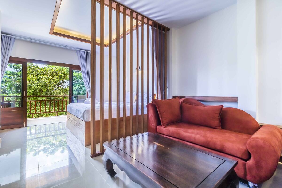 Quarto de hotel moderno com sofá vermelho, mesa de centro de madeira e varanda com vista para o verde.