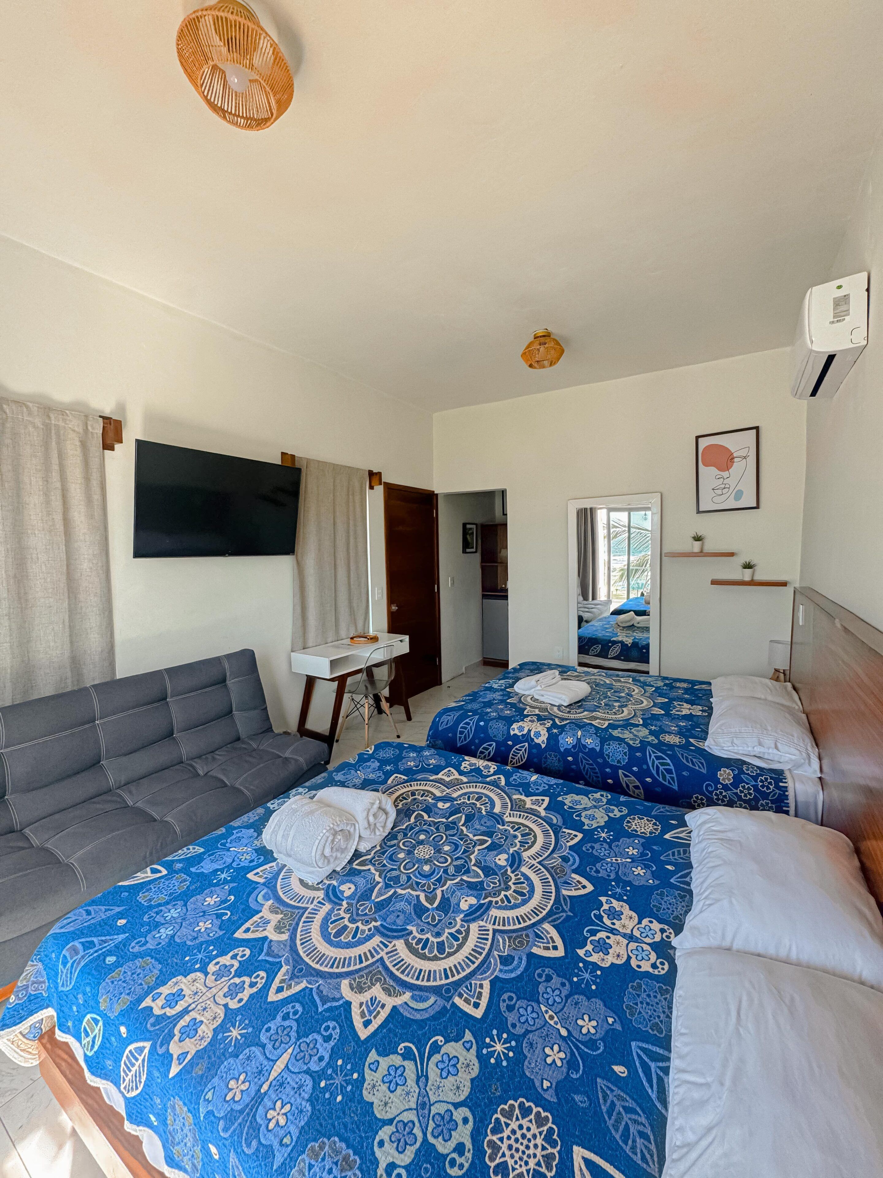 Hotellihuone, jossa on sohva, kaksi sängyä kuviollisilla vuodevaatteilla, televisio, työpöytä ja pieni ruukkukasvi.