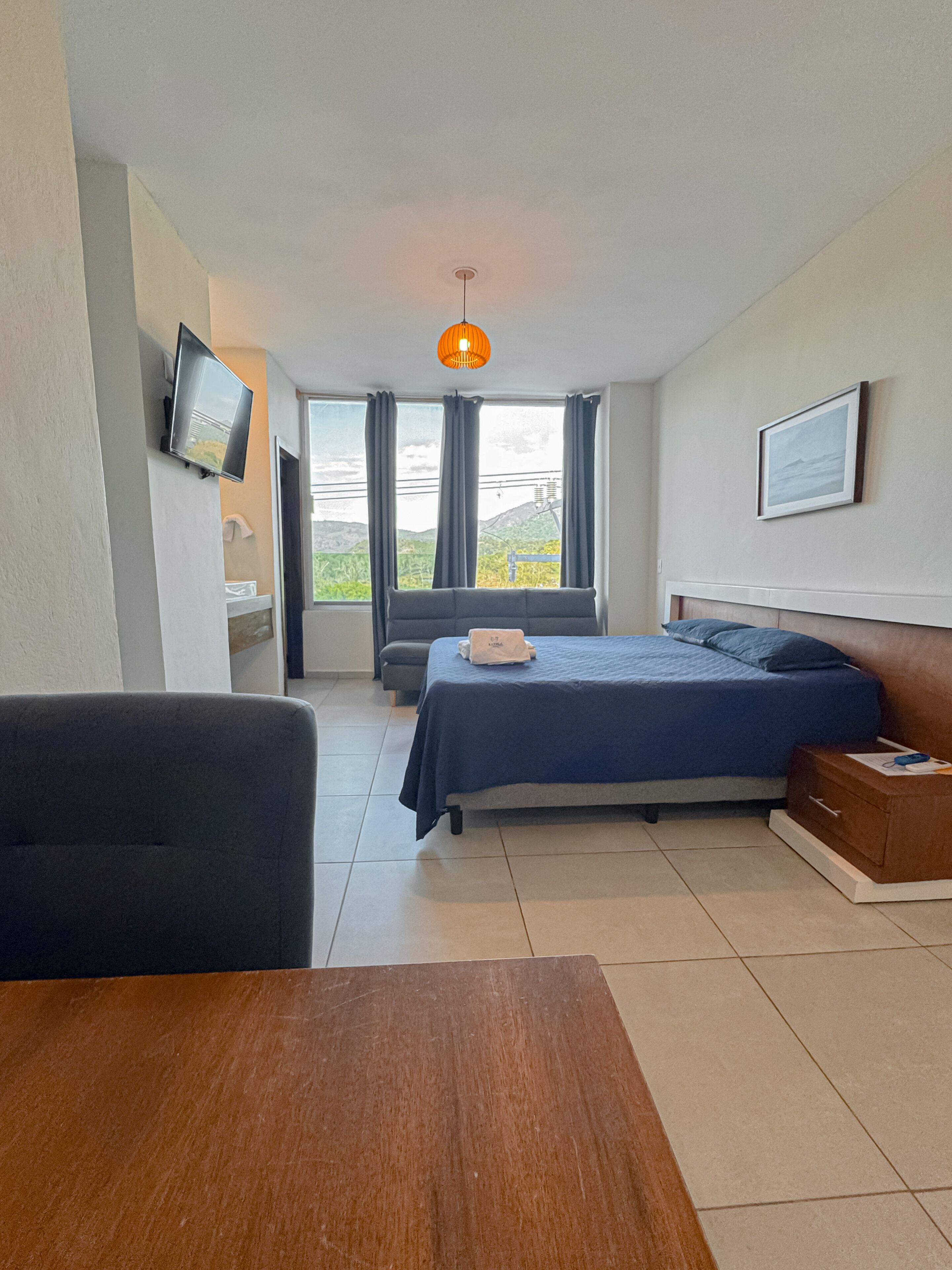 A modern hotel room with a bed, a TV, a wooden table, and a view of the outdoors.