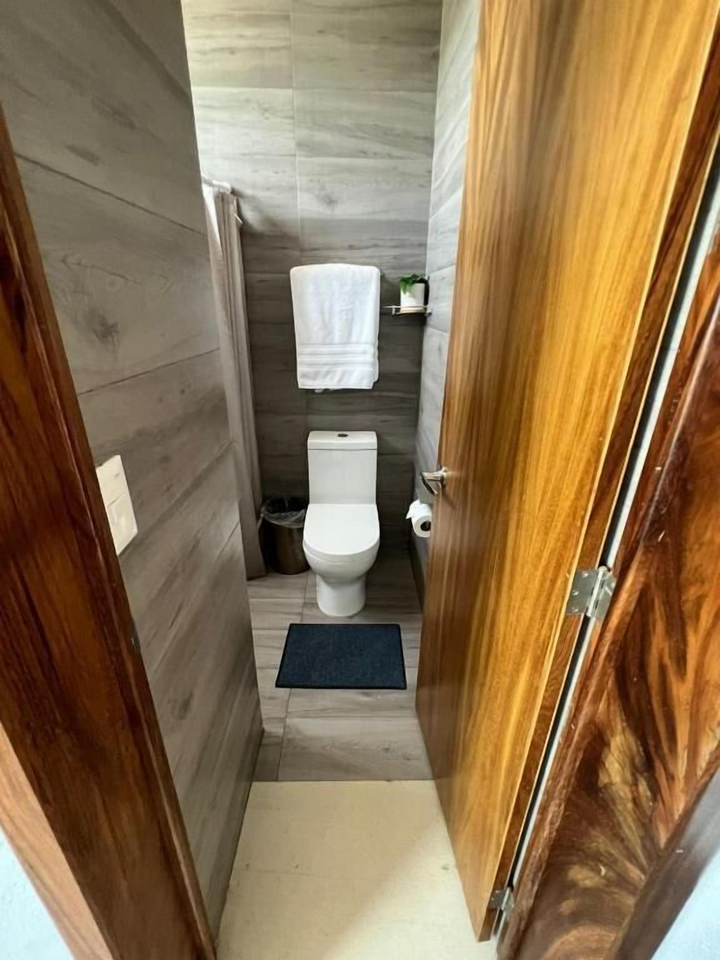 Een smalle badkamer met een toilet, een klein prullenbakje, een handdoek aan de muur en een houten deur.
