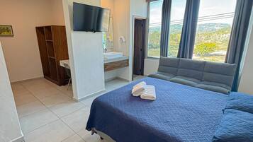 Habitación de hotel con cama, televisor montado en la pared, baño con lavamanos y espejo, y ventana con cortinas.
