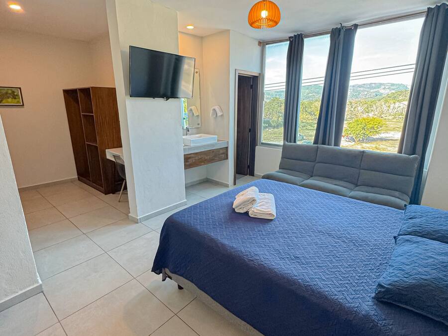 Habitación de hotel con cama, televisor montado en la pared, baño con lavamanos y espejo, y ventana con cortinas.
