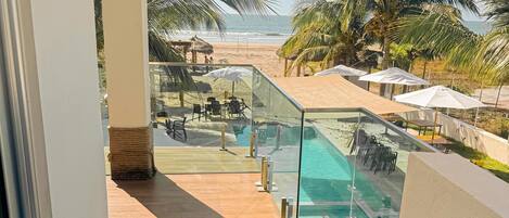 A balcony with a glass railing overlooking a pool area, palm trees, and a beach.