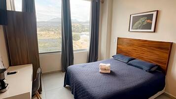 A hotel room with a bed, a desk, a chair, a window with curtains, a ceiling light, and a framed picture on the wall.