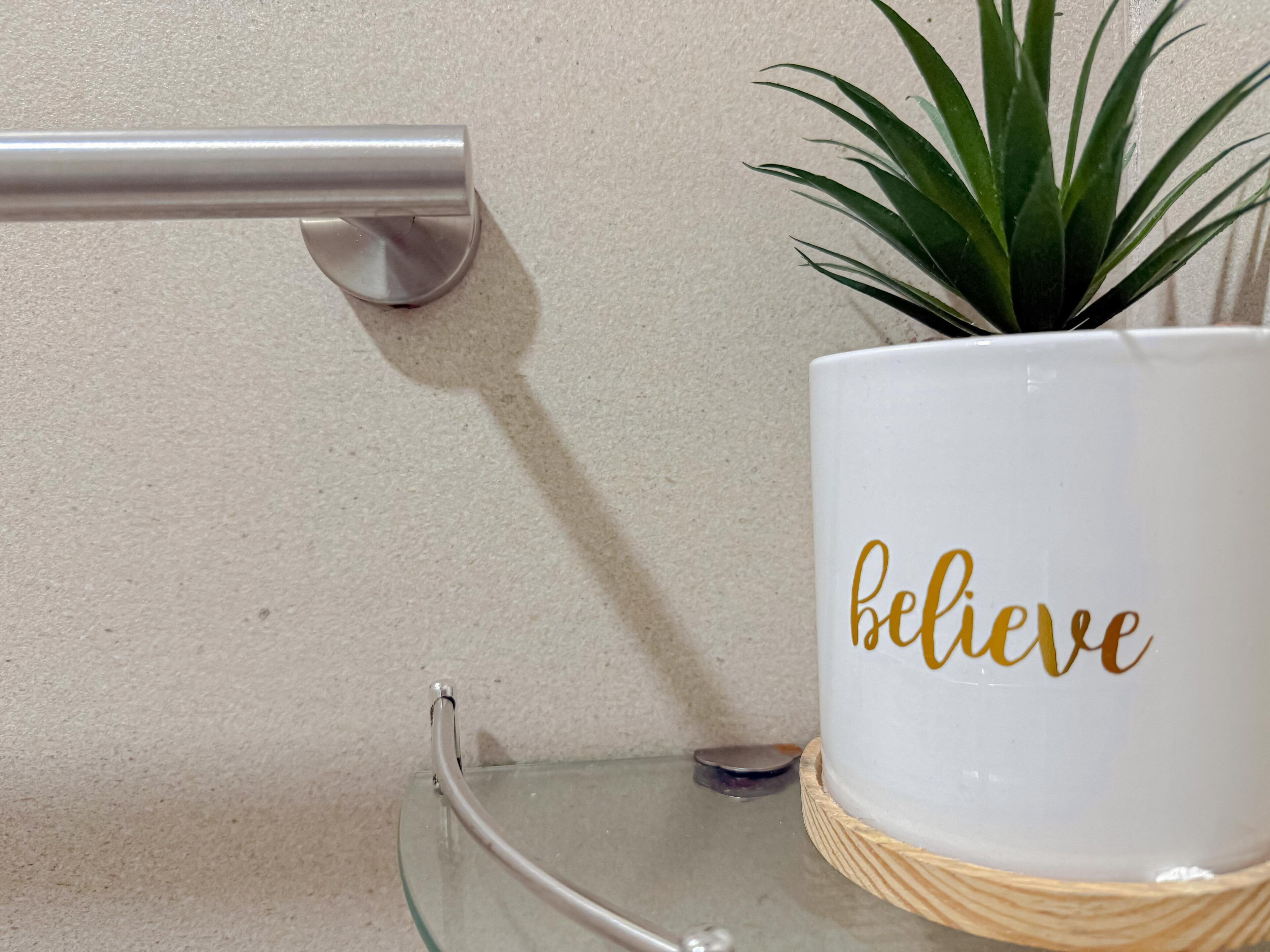 A white mug with the word "believe" in gold lettering, placed on a glass table with a woven coaster.