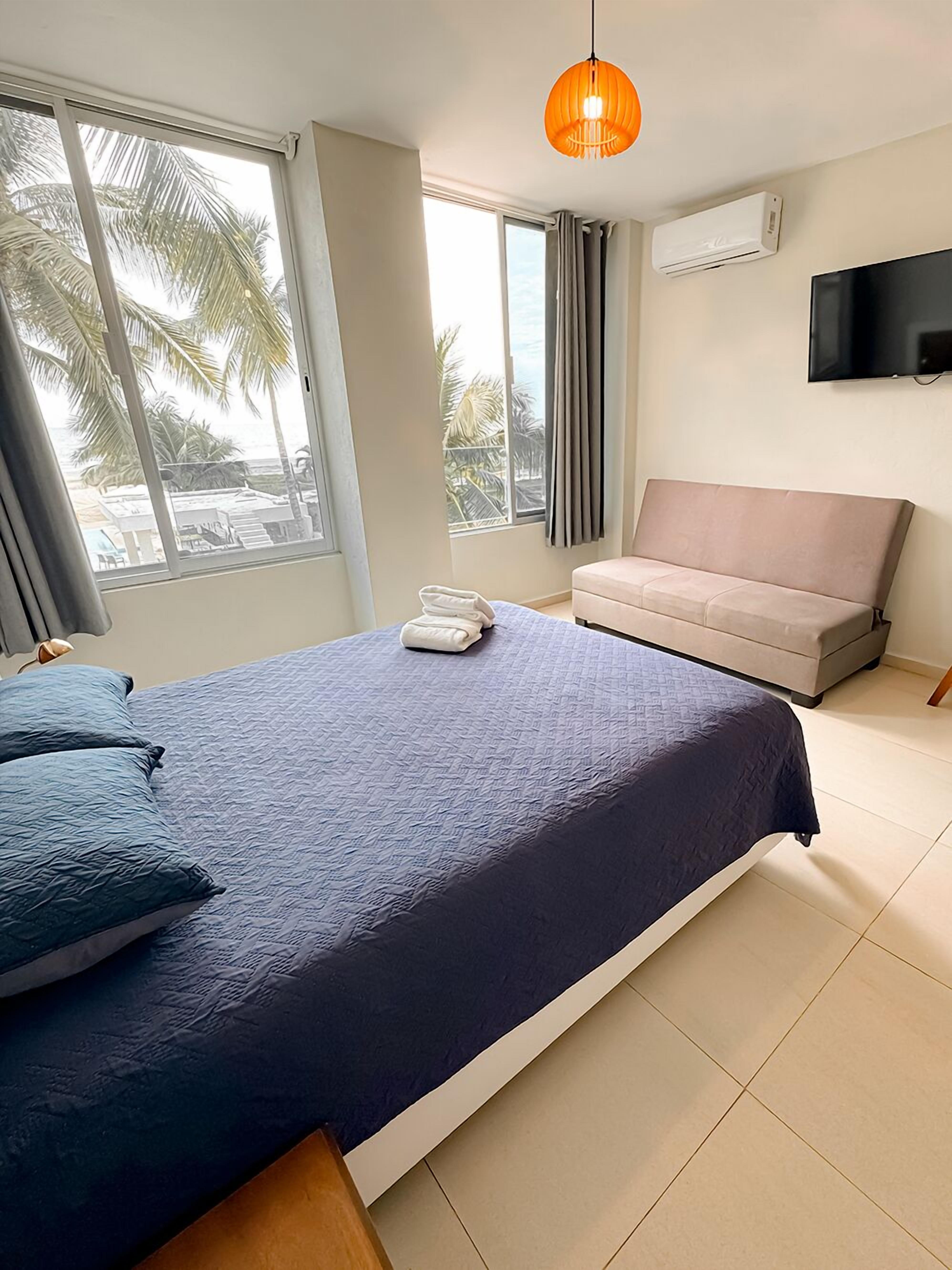 A bedroom with a bed, a sofa, a TV, and a window with a view of palm trees.