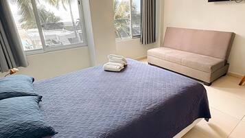 A bedroom with a bed, a sofa, a TV, and a window with a view of palm trees.