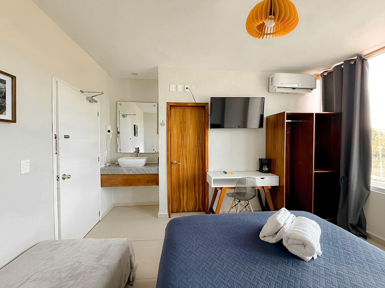 Un dormitorio con cama, escritorio, silla, televisor y baño con espejo y lavabo.