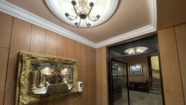 Un couloir aux murs lambrissés, un grand miroir décoratif, un lustre et une porte en verre donnant sur une autre pièce.