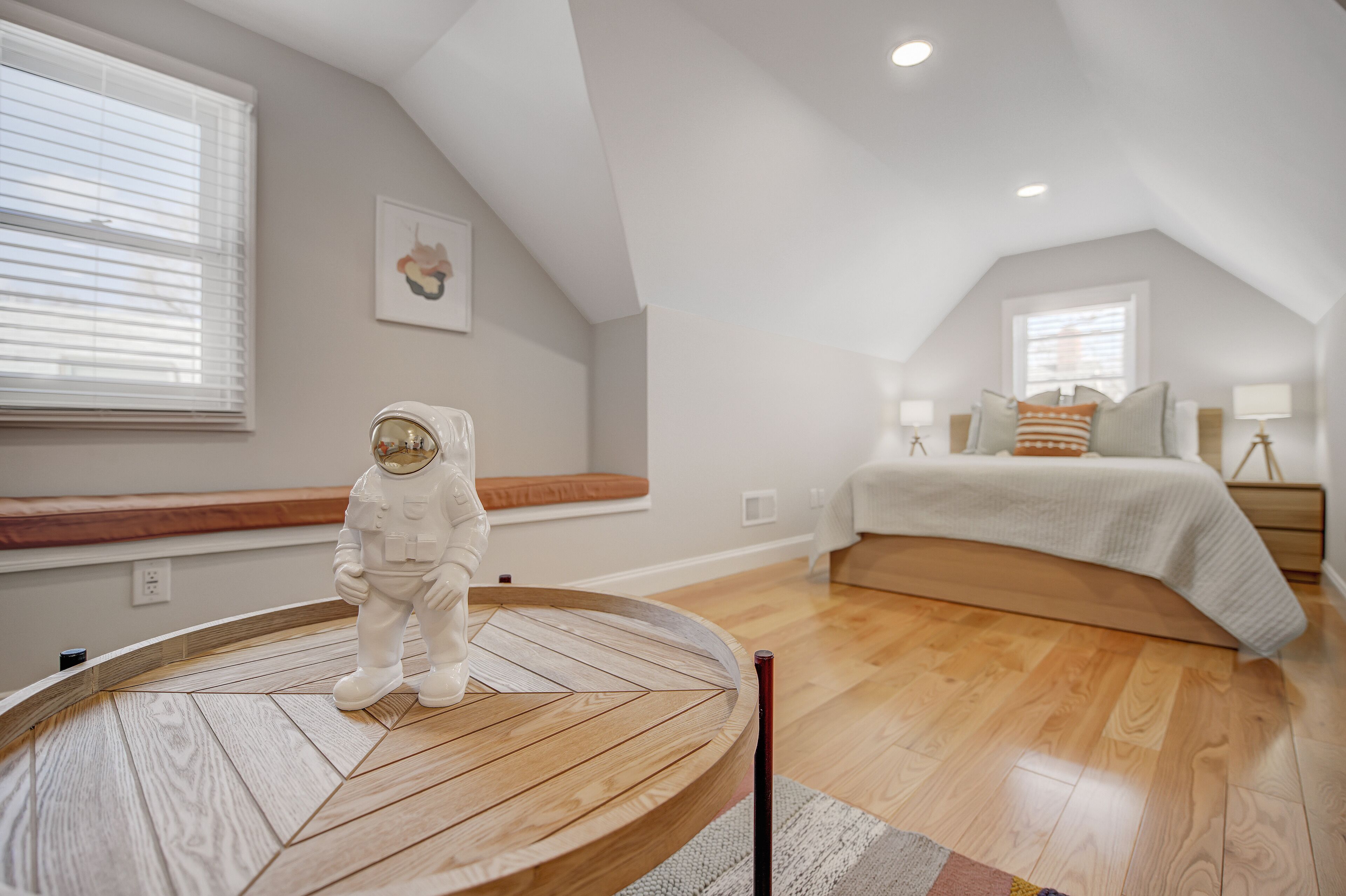 Un dormitorio con techo inclinado, una mesa redonda de madera, una cama con cabecera blanca y una ventana con persianas.