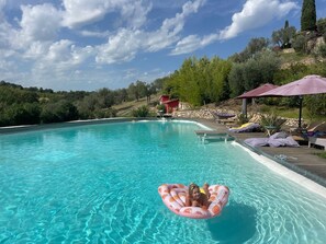 Ein Schwimmbecken mit einer Person auf einem Schwimmschlauch, umgeben von Liegestühlen und Sonnenschirmen, mit einer Hügelkette und Bäumen im Hintergrund.