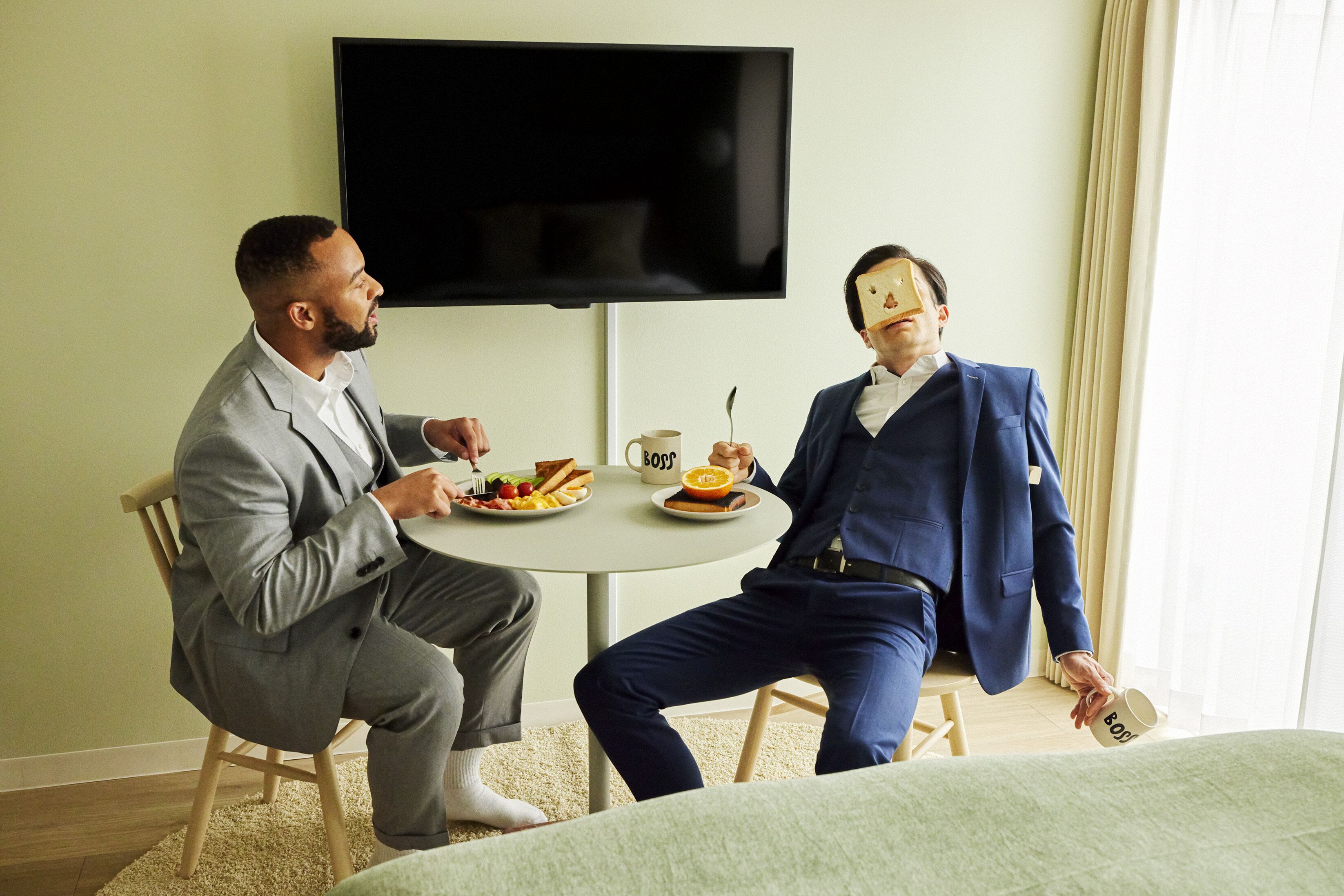Deux hommes sont assis autour d’une table ronde, devant des assiettes de nourriture et des tasses de café, dans une pièce où un téléviseur est fixé au mur et où une fenêtre est ornée de rideaux.