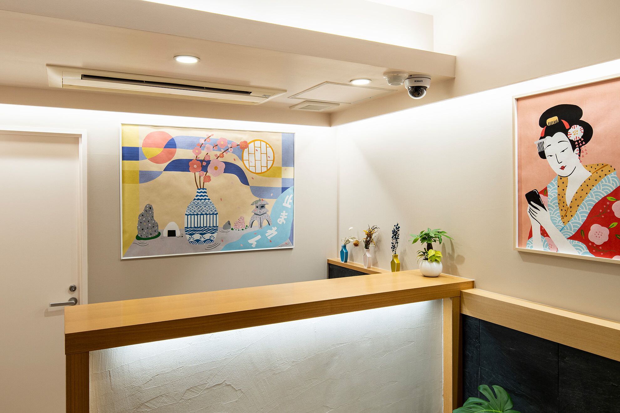 A reception area with a wooden counter, a ceiling-mounted security camera, and two artworks on the wall.