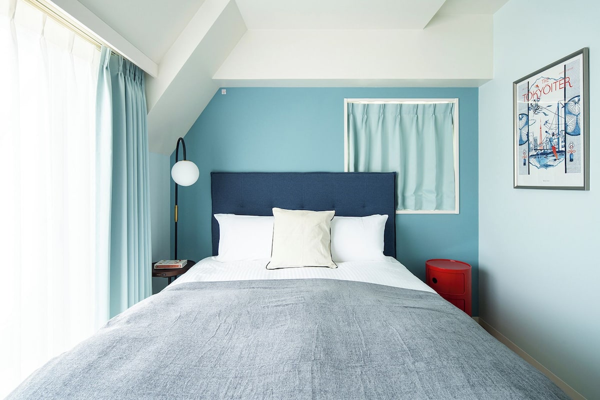 A bedroom with a bed, a nightstand, a red trash bin, a floor lamp, and a wall art piece.