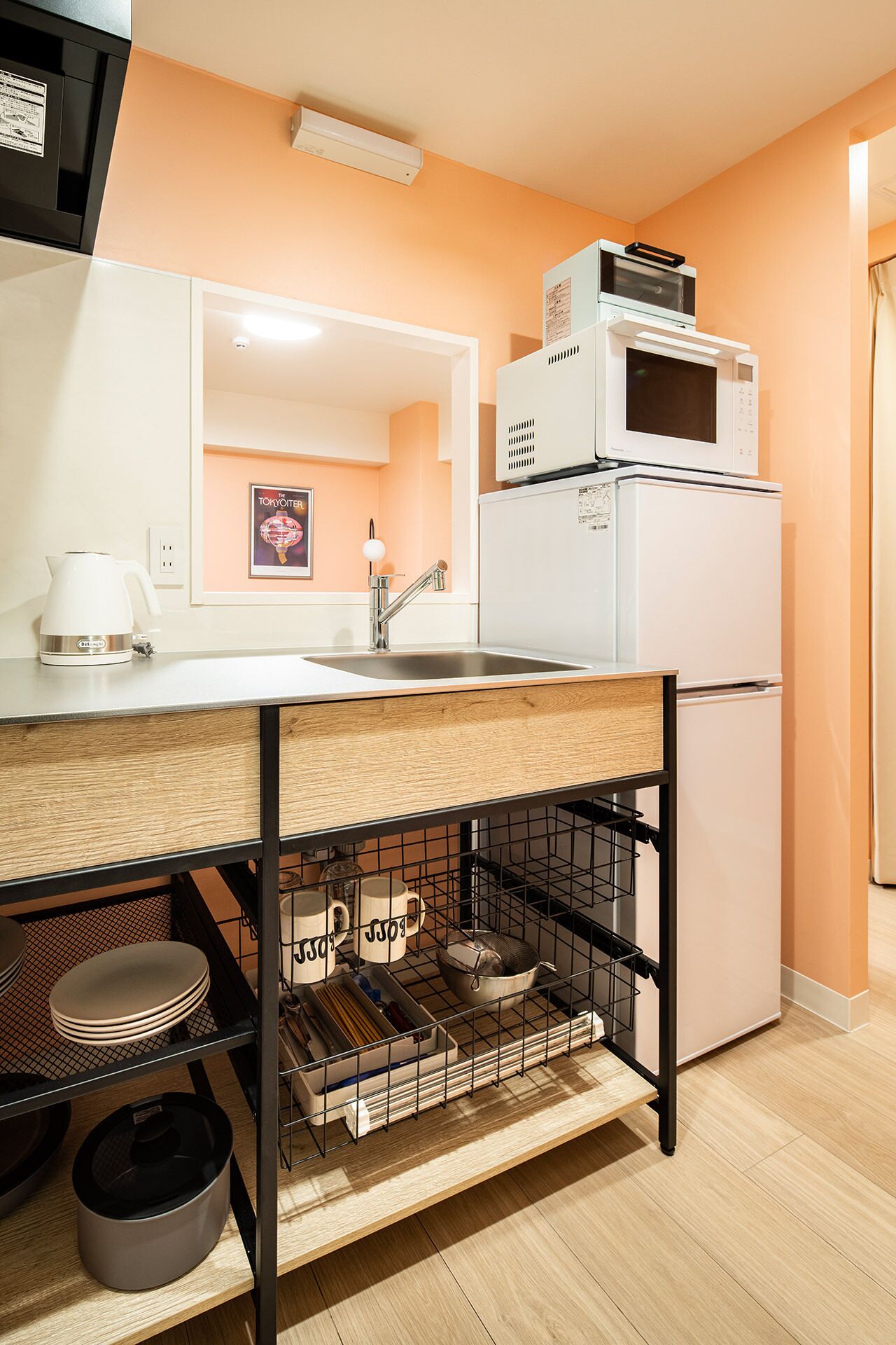 A compact kitchen with a white refrigerator, a microwave on top, a sink with a faucet, and a countertop with a kettle. There is a wooden shelf below holding dishes and a kettle.