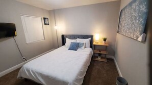Une chambre à coucher avec un lit, une table de chevet avec une lampe, un téléviseur fixé au mur et une fenêtre avec des stores.