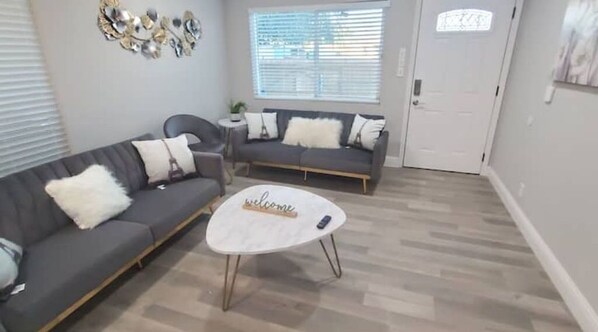 A living room with a grey sofa, a round coffee table with a 'welcome' sign, and a small side table with a plant.