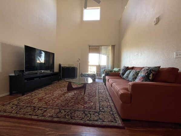 A living room with a flat-screen TV, a glass coffee table, a patterned rug, a sofa, and a sliding glass door leading to a balcony. - Biltmore Area Spacious 2 bedrooms 2 baths townhome with a bonus room and balcony (Phoenix)