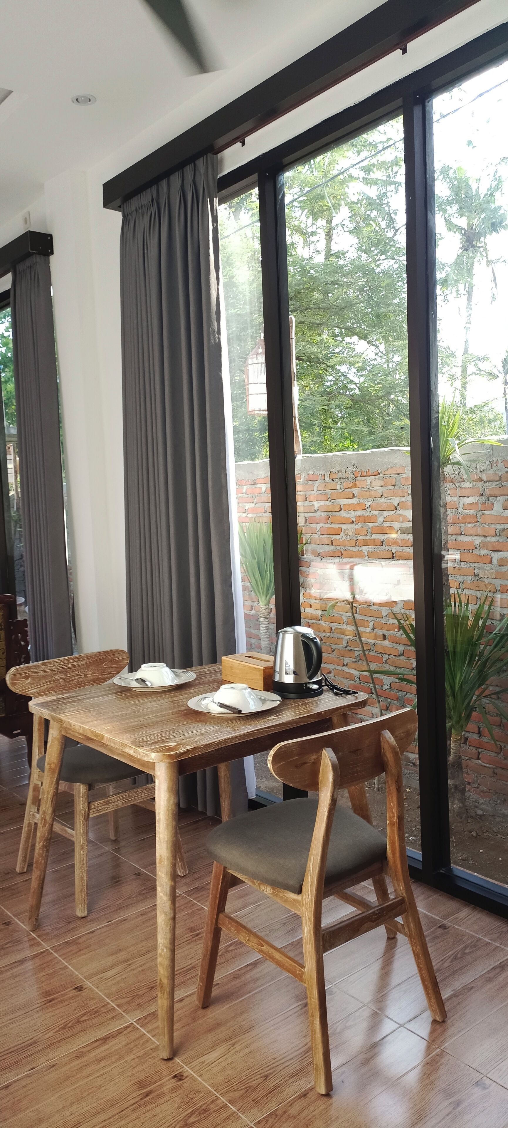 A dining area with a wooden table and chairs, a kettle, and a brick wall visible through the window.