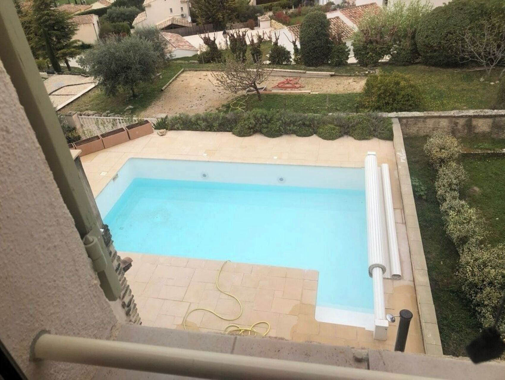 Una piscina rectangular con una solana empaveda alrededor, ubicada en un jardín ajardinado con diversas plantas y vistas a casas vecinas.