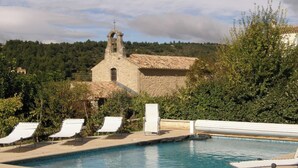 Una piscina con sillas de descanso, un edificio de piedra con una torre de campanas y un paisaje boscoso al fondo.