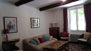 Una sala de estar con sofá, mesa y una silla. Hay dos cuadros en la pared, una lámpara y una ventana con cortinas.
