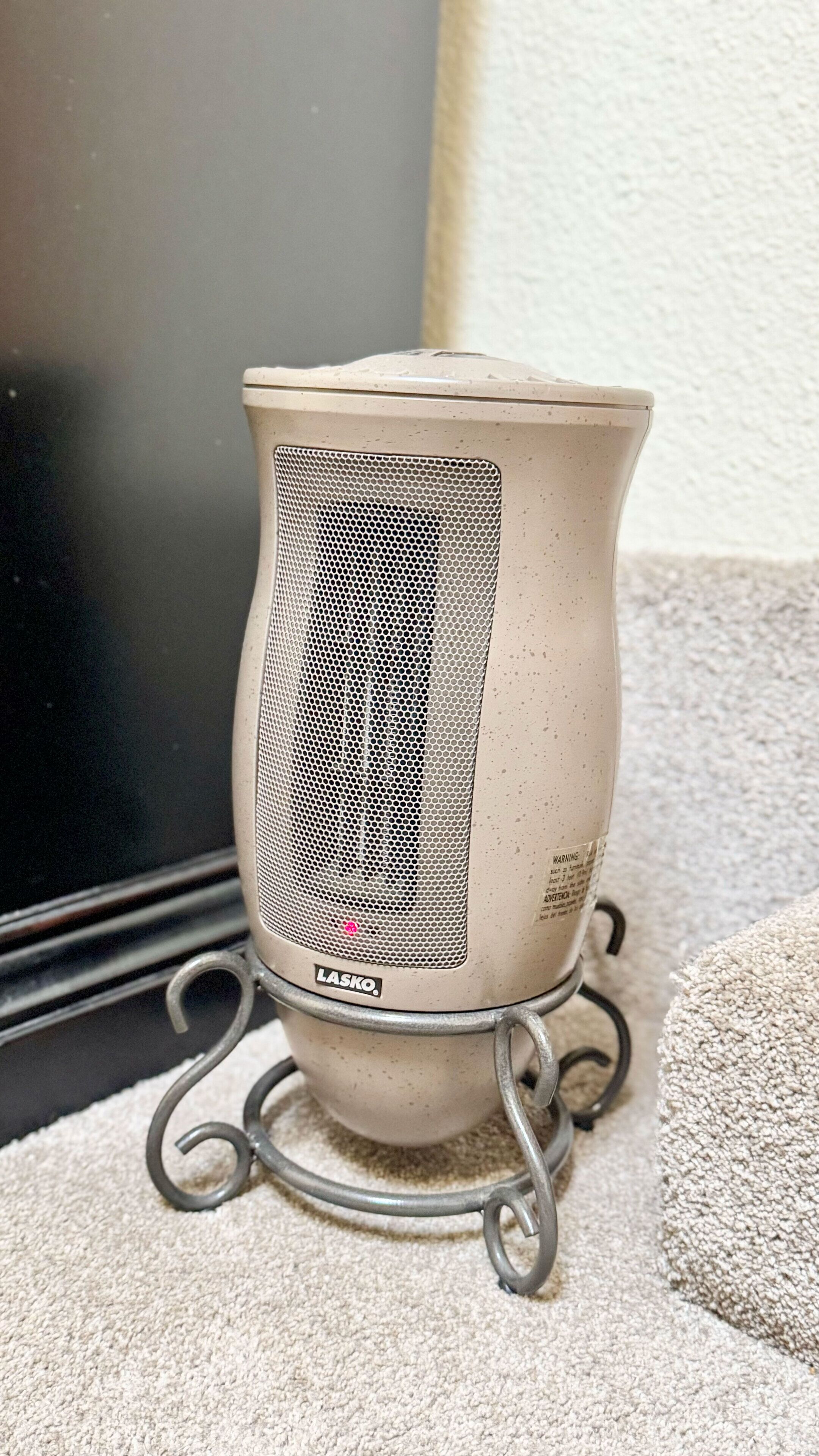 A portable heater with a mesh front, standing on a metal stand, placed on a carpeted floor.