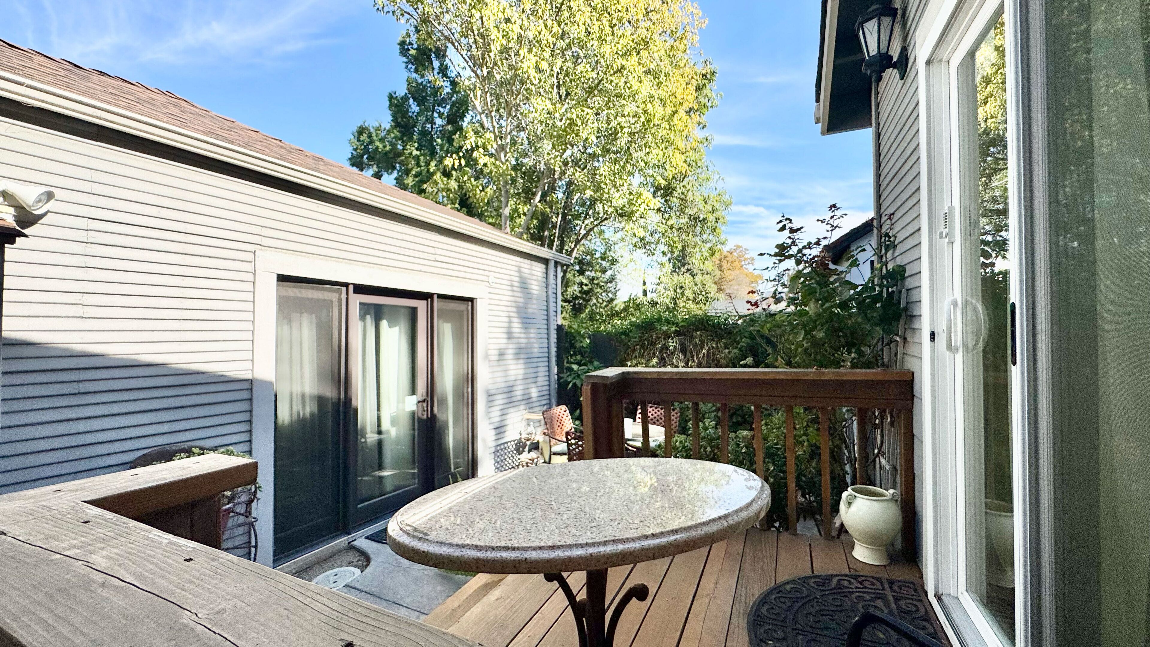 Une terrasse en bois avec une table ronde et des chaises, un petit coin jardin agrémenté d’une plante en pot, et une porte-fenêtre coulissante donnant sur un patio.
