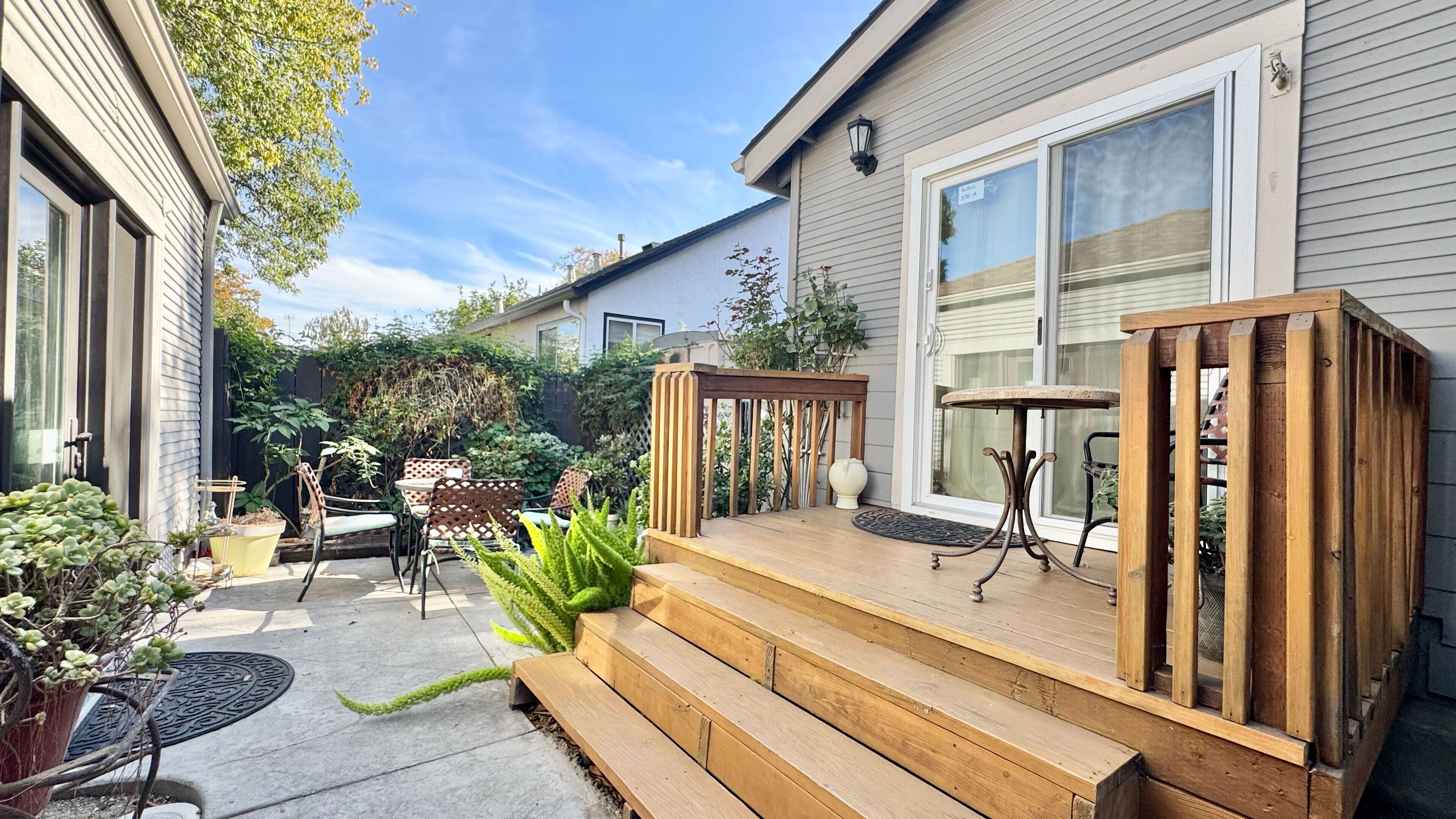 Een houten terras met een bank, een tafel en stoelen. Er staat een plant in een pot en een kleine tuin met diverse beplanting.