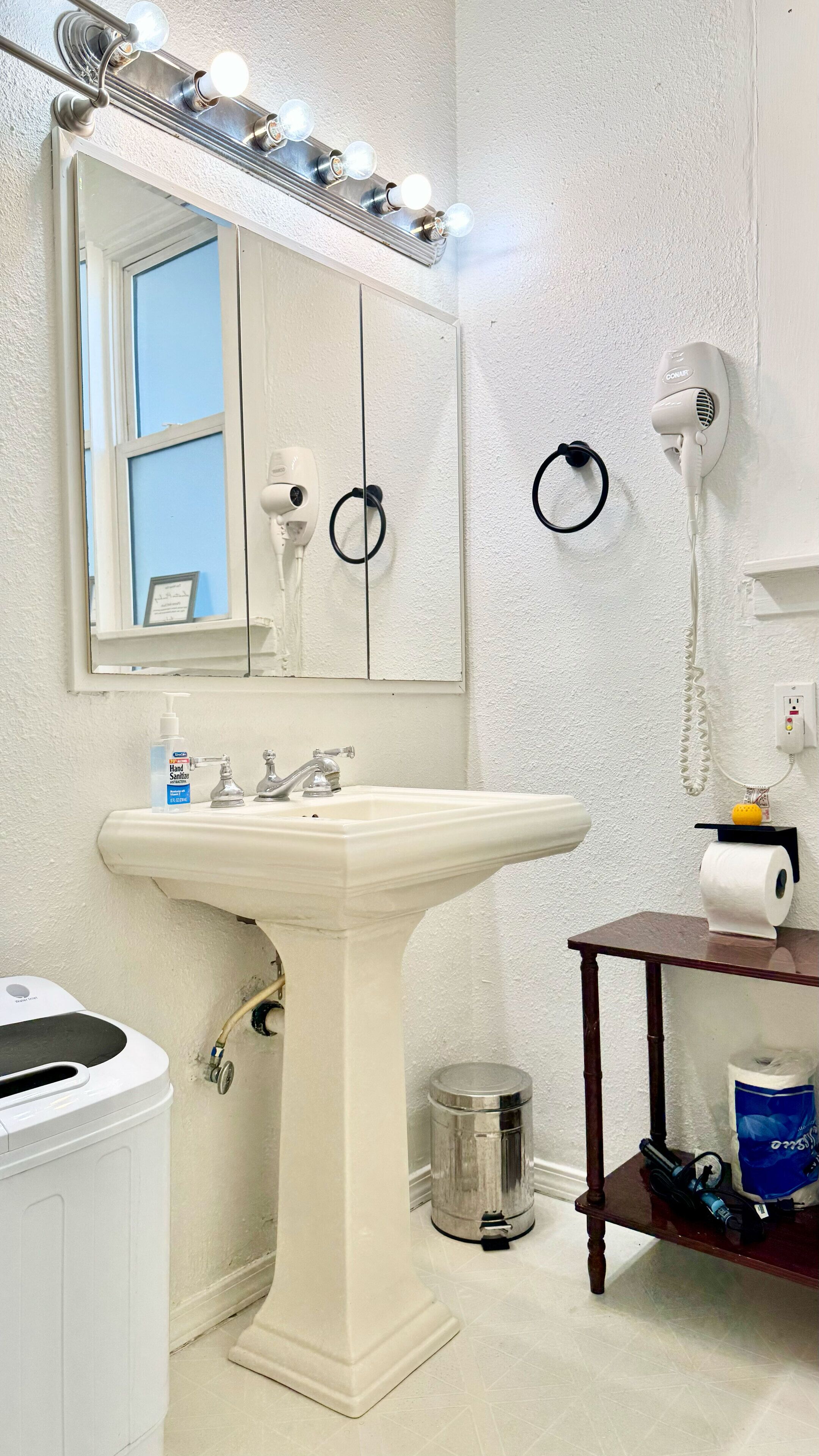 Een badkamer met een wit wastafel, een spiegel met verlichting, een haardroger en een handdesinfecterend middel.