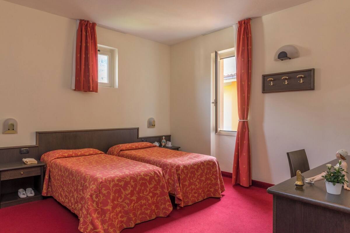 Een hotelkamer met twee bedden, een bureau, een stoel, een klein tafeltje, een raam en een wandplank.