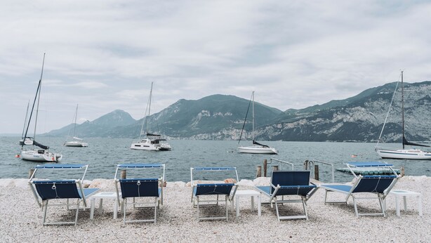 Ein Kiesstrand mit Reihen blauer und weißer Liegestühle, vor einem ruhigen See mit Segelbooten und Bergen im Hintergrund.